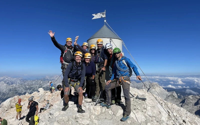 Group of climbers smiling and raising their arms next to the Aljaž Stolp on the summit of Mount Triglav