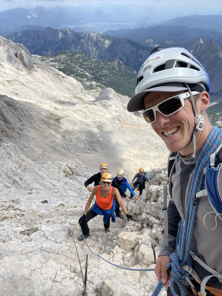 Smiling mountain guide wearing a helmet and sunglasses leading climbers on rocky terrain toward Mount Triglav