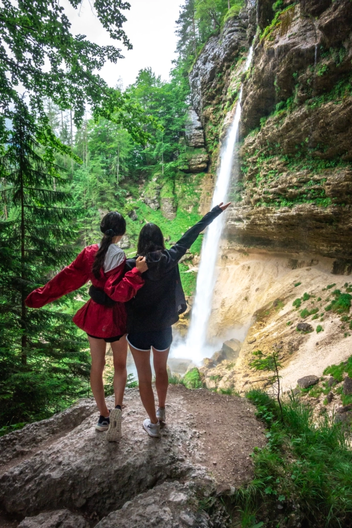 Hikers walking behind a waterfall on a rocky trail surrounded by green forest